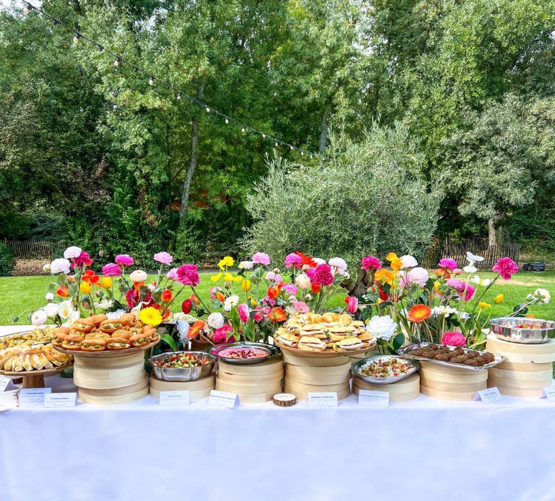 Table de buffet Entre L dressée dans un jardin : mezzés, fricassés, petits pains et falafels entre des fleurs